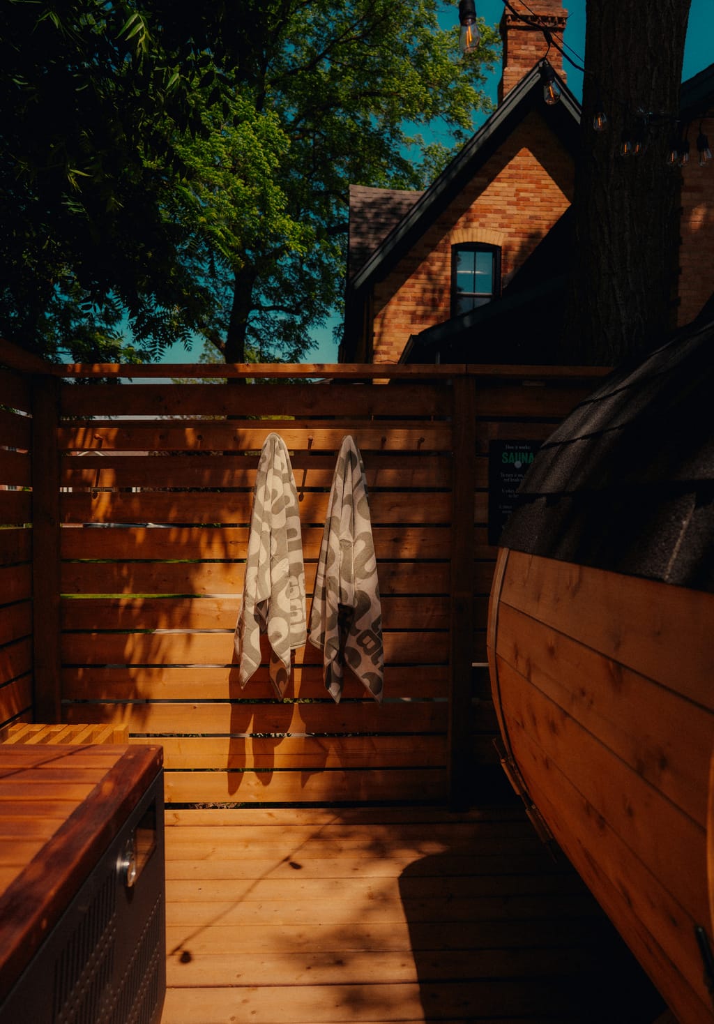 Barrel sauna and two towels hanging on a cedar fence in a backyard, brick house behind the trees