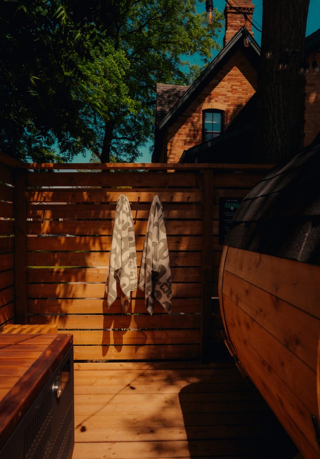 Barrel sauna and two towels hanging on a cedar fence in a backyard, brick house behind the trees