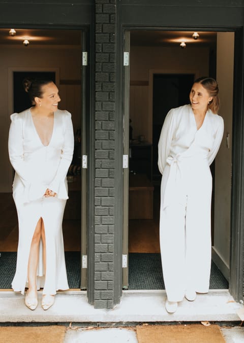 Two women in white outfits smile at each other from neighbouring doorways in a black brick facade