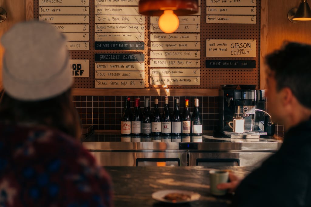 Two guests at the bar facing a chalk menu board, a row of wine bottles and an espresso machine
