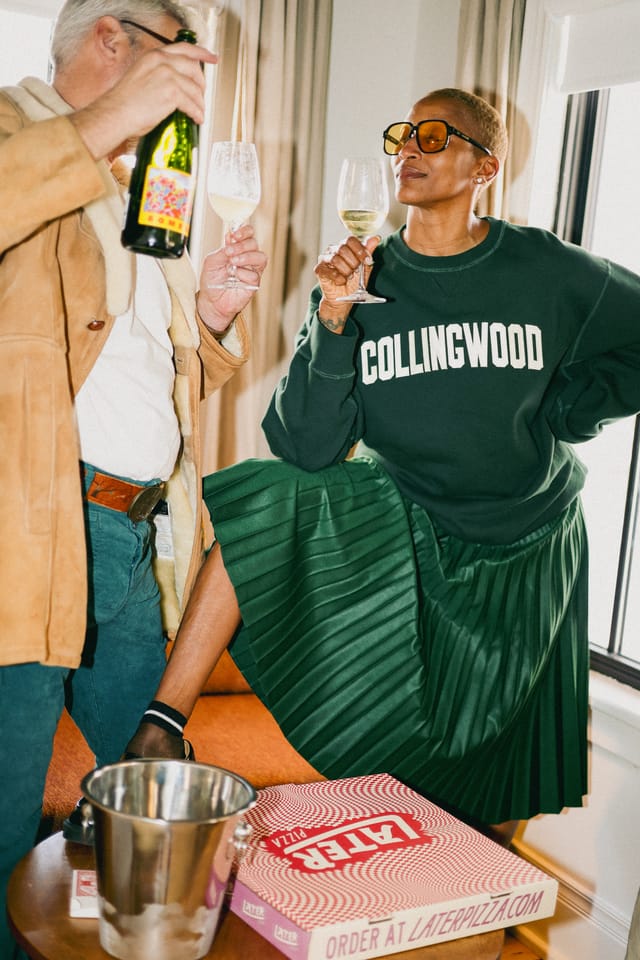 Two people raising glasses of white wine and a bottle in a room, a pizza box on the side table