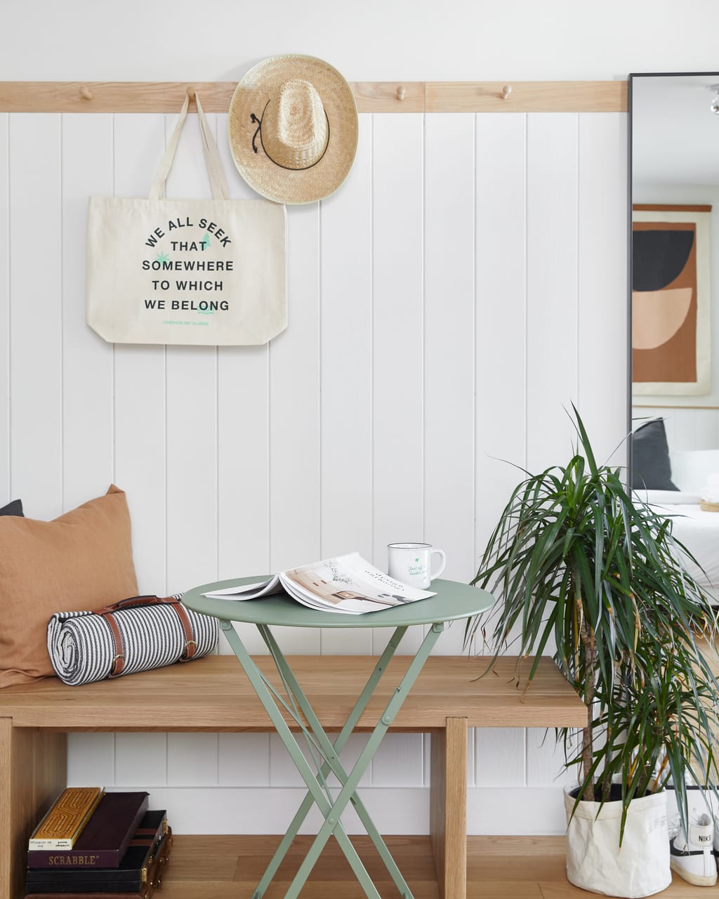 Wall pegs with a tote bag and straw hat above a bench and green bistro table with a magazine