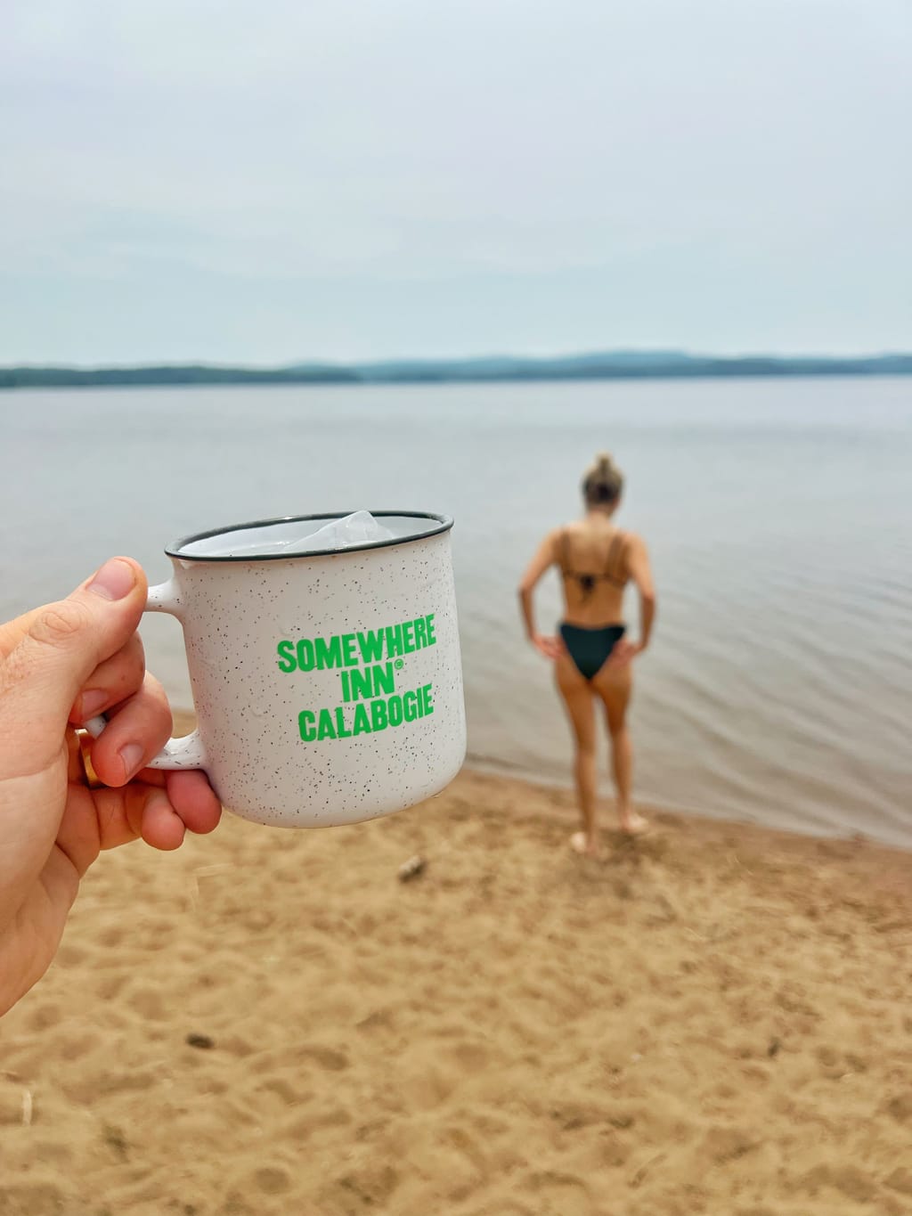 Somewhere Inn Calabogie mug held up on a sandy beach, with a swimmer at the lake edge