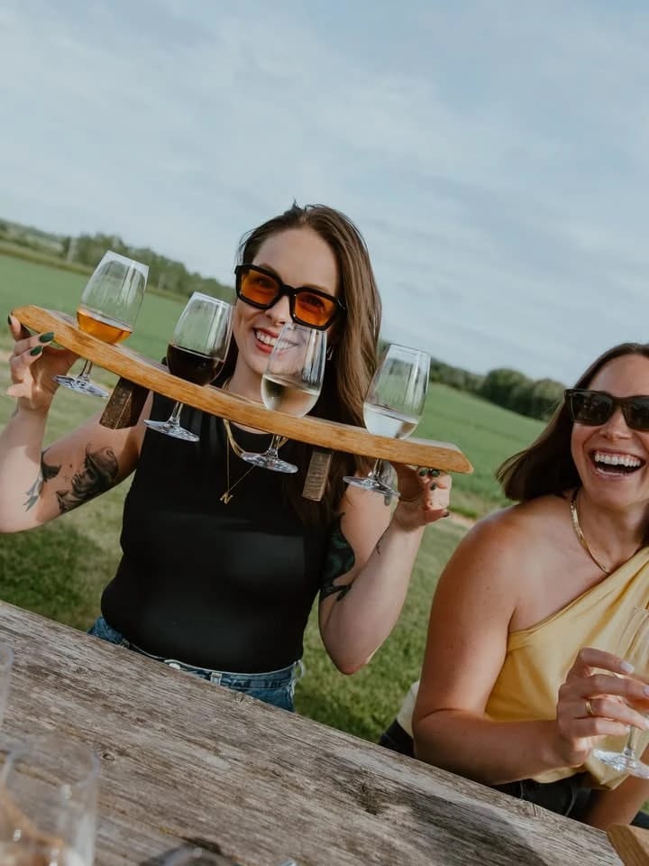 Two women laugh at a picnic table, one holding a wooden flight paddle of four wines, a field behind