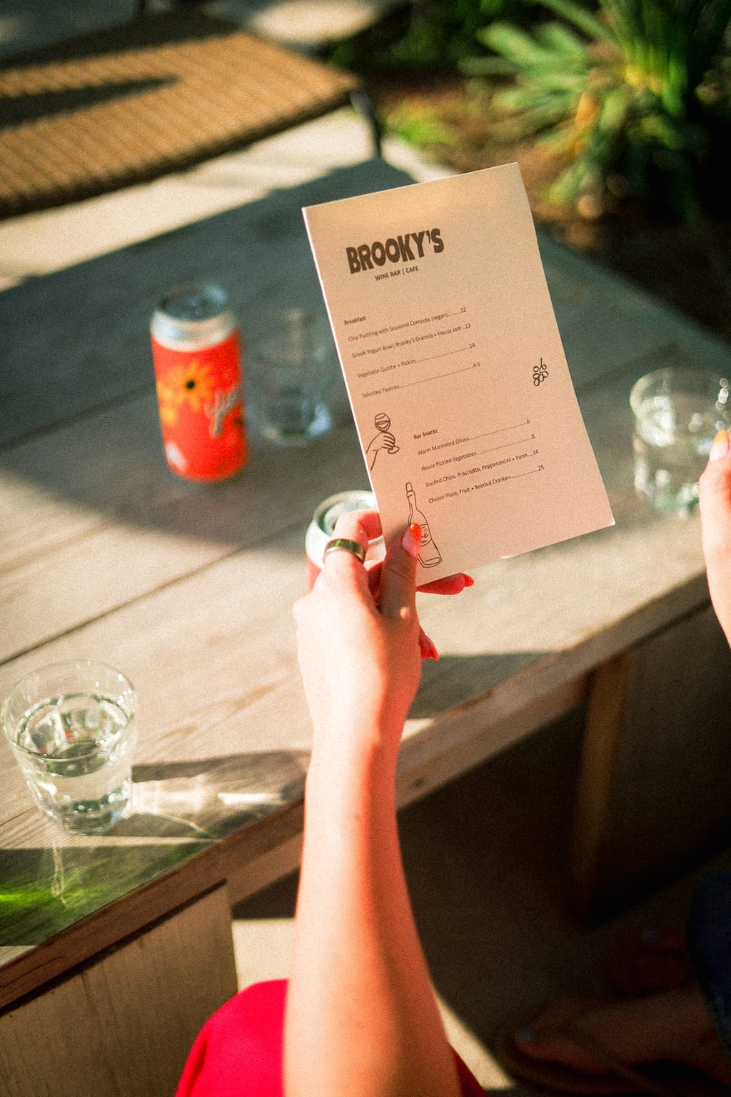 Hand holding a Brooky's Wine Bar and Cafe menu above a patio table with a can and glasses