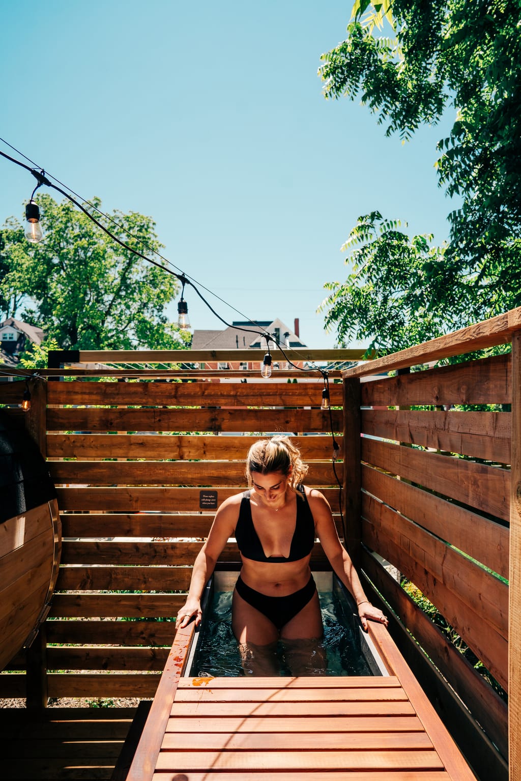 Woman stepping into the cold plunge tub in a cedar-fenced garden under string lights