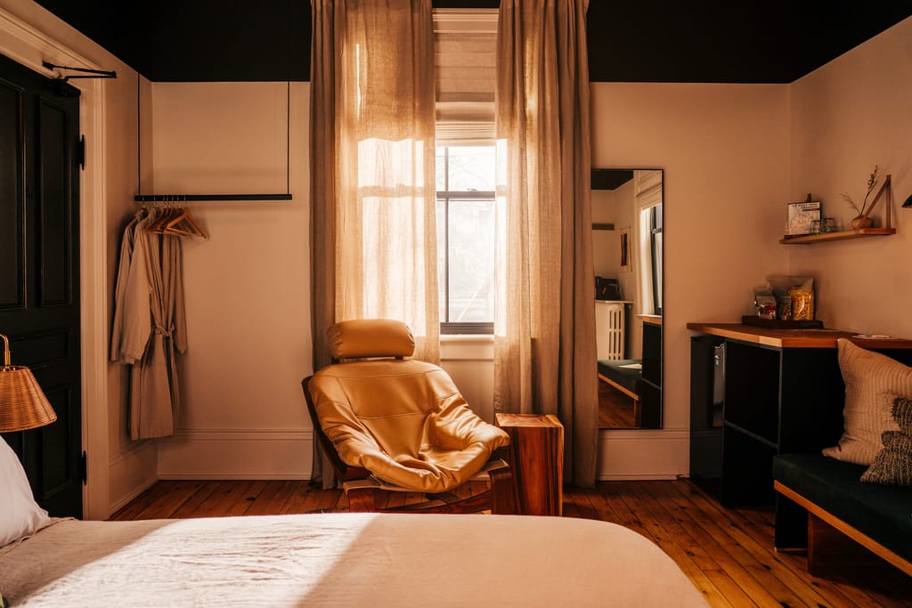 Guest room with linen curtains, a leather lounge chair, a mirror, robes on a rail and pine floors