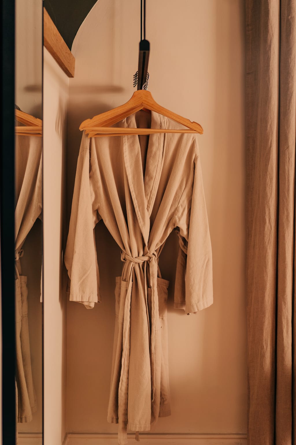 Linen robe hanging on a wooden hanger beside a mirror and a linen curtain