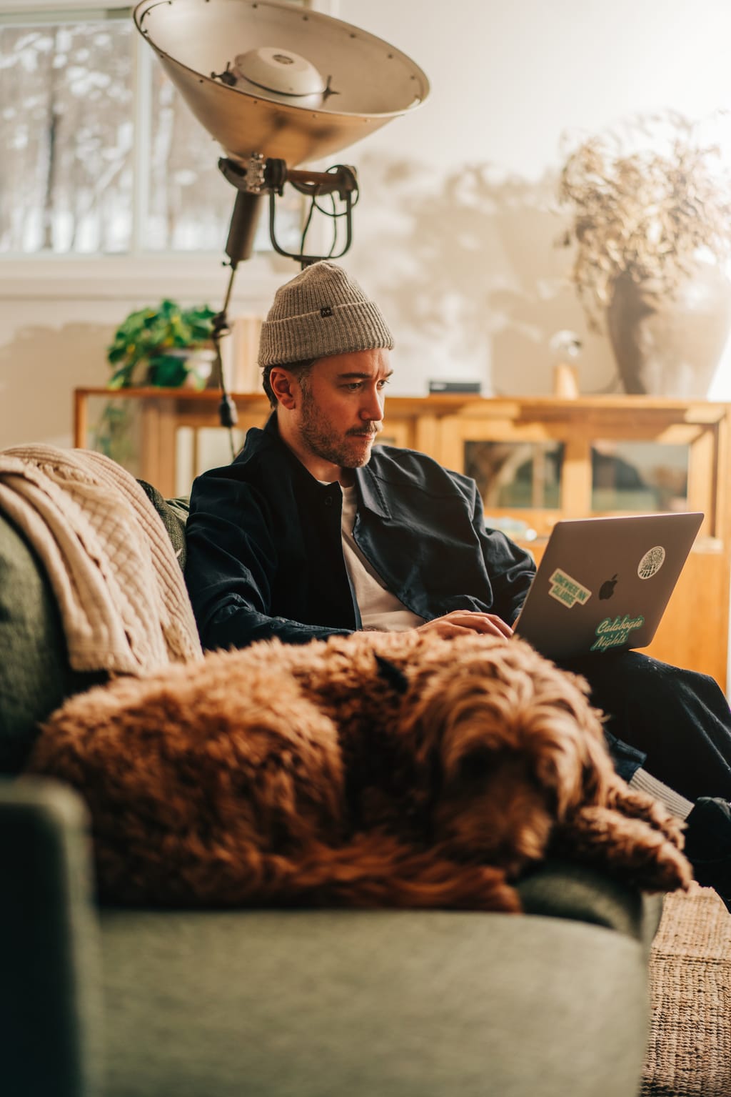 Man in a beanie working on a laptop on a green sofa, a curly brown dog asleep in front