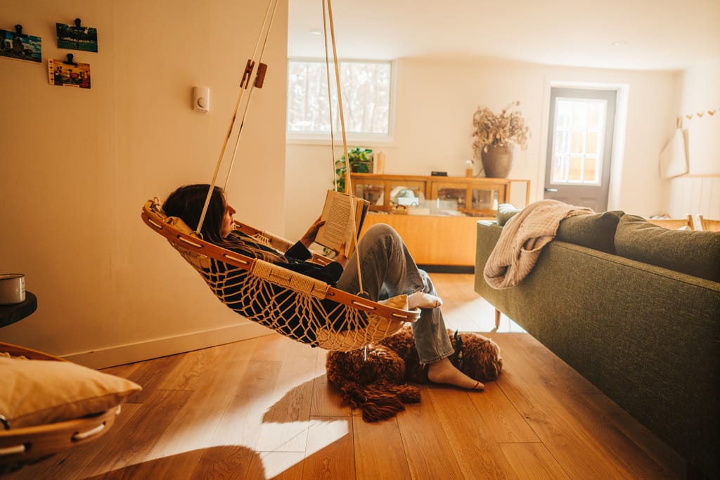 Woman reads in a hanging rope chair beside a green sofa, a dog curled on the wood floor