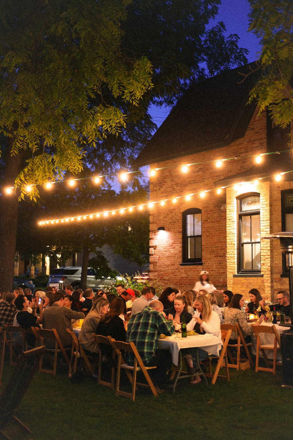 Guests dining at long tables under string lights in a garden beside a brick house at night