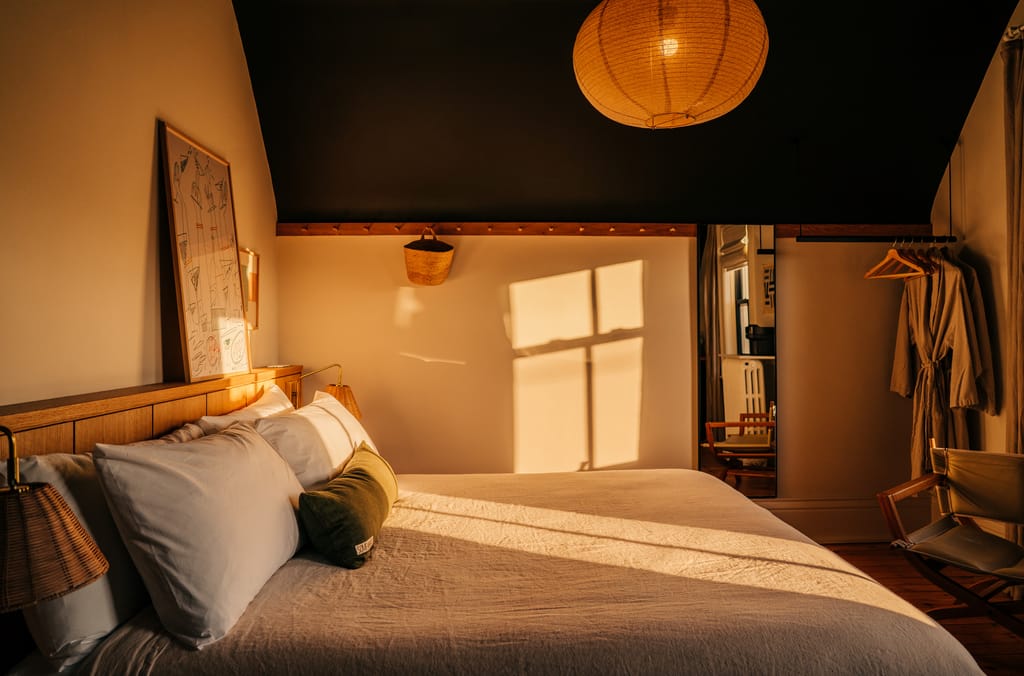 Bed with white linens and a green bolster in an attic room with a paper lantern and robes on a rail