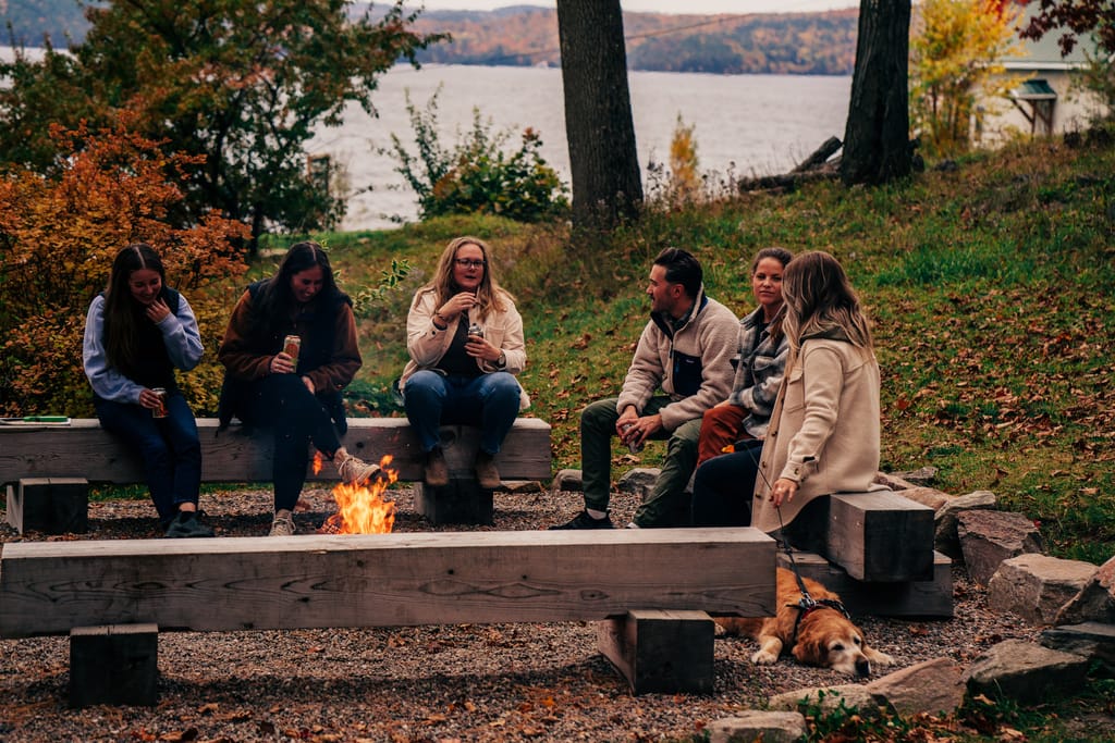Friends sitting around a fire pit with a dog, the lake and autumn trees behind them