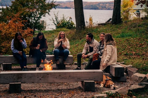 Friends sitting around a fire pit with a dog, the lake and autumn trees behind them
